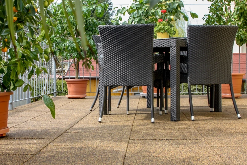Een zitgroep staat op een terras dat is aangelegd met vierkante terrasplaten in een aardkleurig dessin. Langs de gegalvaniseerde reling staan citrusbomen.