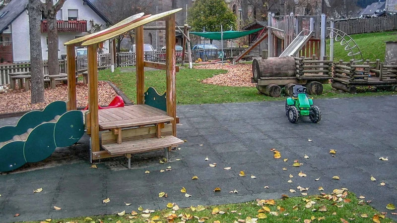 Blick auf den zentralen Bereich eines abwechslungsreich und naturnah gestalteten Spielplatzes in Olsberg. Der Spielplatz besteht im Zentrum aus einer großzügig angelegten Spielfläche aus grünen Fallschutzplatten, auf der ein kleines Spielhaus steht.