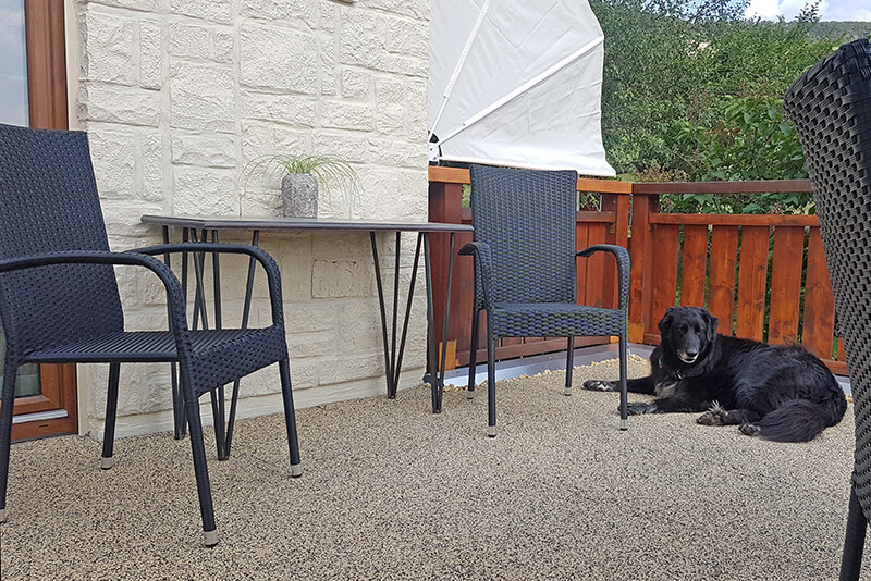 Zwarte hond liggend op balkon met grijze balkontegels. Twee stoelen en een tafel staan bij het huis. Over een bruine houten reling gaat het uitzicht naar groene bomen.