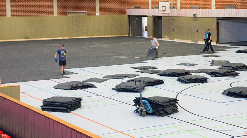 In een grote sporthal wordt een evenementenvloer gelegd. Deze bestaat uit rubbergranulaatmatten in een in elkaar grijpend bestratingsontwerp. De evenementenvloer beschermt de gevoelige sportvloer van de hal tegen beschadigingen.