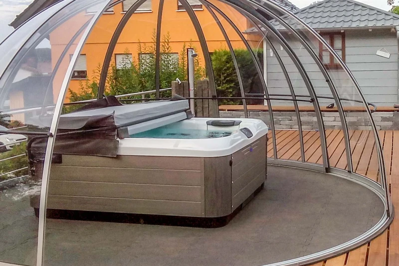 Een halfopen Glasiglo met whirlpool staat op een houten terras voor een vrijstaand huis. De vloer van de Glasiglo bestaat uit waterdoorlatende vloertegels van WARCO.