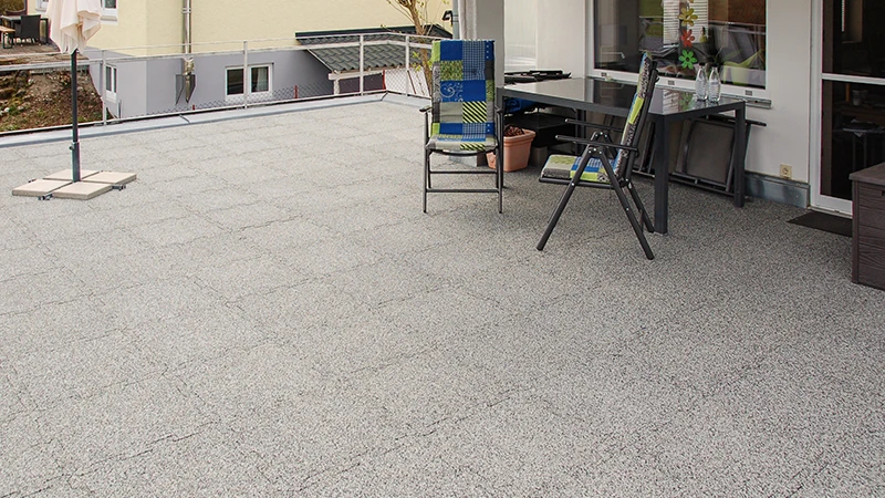 Op een dakterras liggen grijs gemarmerde WARCO-terrastegels van PU-gebonden rubbergranulaat. De puzzelverbinding garandeert een nauwkeurige pasvorm en combineert weerbestendigheid met duurzaamheid. Het vormstabiele materiaal dempt voetstappen en zorgt voor een opgeruimde, verzorgde uitstraling. Bovendien biedt het een slipvaste ondergrond en hoog loopcomfort, wat de vloer extra gebruiksvriendelijk maakt.