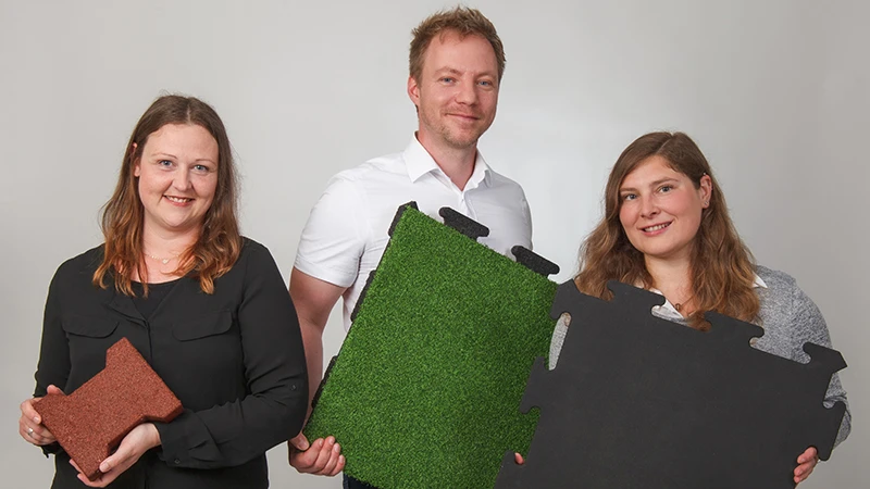 Met een vriendelijke glimlach tonen drie adviseurs van WARCO diverse vloertegels van PU-gebonden rubbergranulaat in bruin, groen en zwart. Deze tegels zijn speciaal geschikt voor balkons en bieden een slipvaste, duurzame oplossing. Elke tegel heeft een eigen ontwerp en is eenvoudig te plaatsen. Dankzij hun deskundige begeleiding vind je de juiste vloerbedekking voor een veilig en stijlvol balkon. De onderhoudsarme tegels zorgen bovendien voor extra comfort.