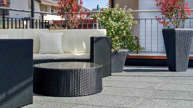 Op een zonovergoten dakterras in een moderne nieuwbouwwijk kijkt men neer op een zithoek gemaakt van rotan meubilair. Op de achtergrond staan plantenbakken, ook gemaakt van polyrattan. De grijze terrasvloer is gemaakt van WARCO terrasvloeren in een licht granietkleurig design.