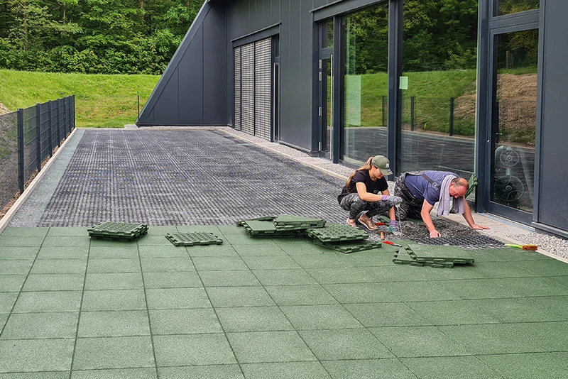 Voor een donkere, moderne gevel installeren een vrouw en een man groene WARCO-terrastegels van PU-gebonden rubbergranulaat. De tegels sluiten naadloos op elkaar aan en vormen een slipvaste, weerbestendige en duurzame toplaag op het terras. Dit stevige materiaal biedt comfort en bescherming bij elk weertype. Dankzij de eenvoudige installatie is een persoonlijke invulling mogelijk en krijgt ieder terras een hoogwaardige, eigentijdse uitstraling.