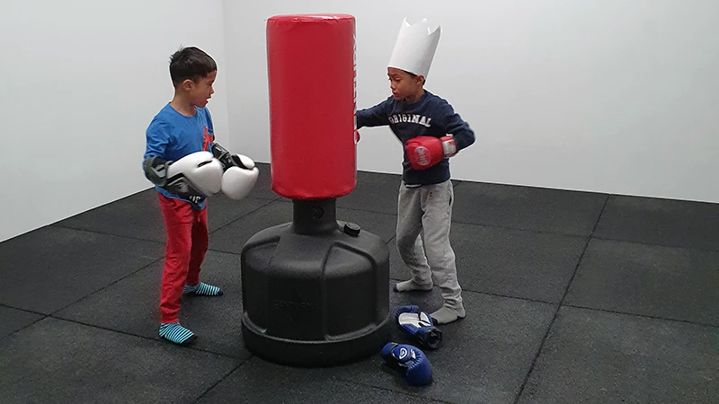 Twee kinderen boksen op één apparaat, een staande bokszak. De vloer is bedekt met zwarte Warco-matten.