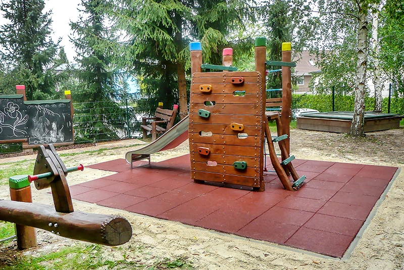 Valhoogte en impactgebied Een bijna natuurlijke speeltuin met houten speeltoestellen ligt op een terrein met bomen. Er zijn planken om op het hek te schilderen. In het midden van de speeltuin staat een kleine houten klimtoren met glijbaan en klimwand. Het botsgebied rond de klimtoren is bedekt met WARCO valbeveiligingsplaten.