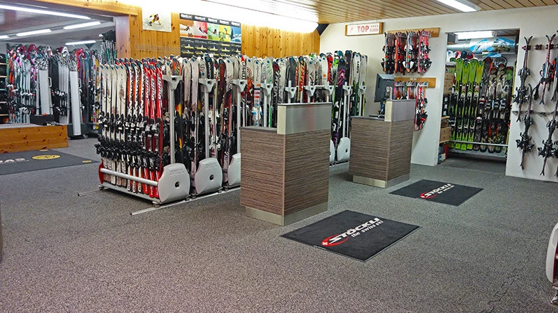 Achter twee balies in de skiwinkel Gaby Sports staan stands met verschillende modellen. De vloer is bedekt met kliktegels binnenshuis van WARCO in de tint rotan lounge.