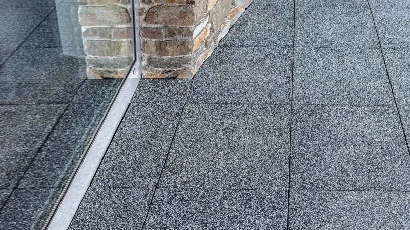 Op een terras liggen vierkante terrasplaten in de kleur designgrijs graniet. De terrasdeur grenst aan een schuine natuurstenen muur van zandsteen. De terrasplaten van WARCO zijn op het profiel van de muur gesneden en liggen vlak zonder voeg.