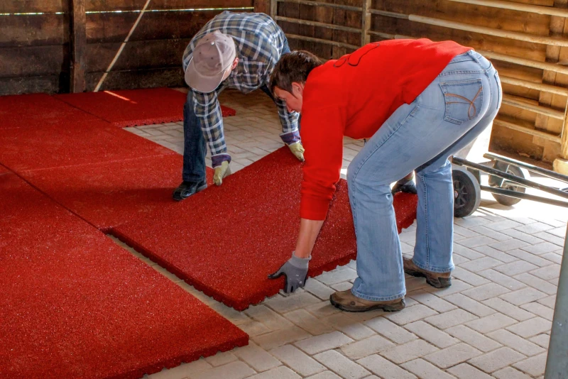 Twee mensen zijn WARCO stalmatten aan het leggen in een paardenstal waarvan de stalvloer al is vastgelegd met betonnen bestrating. Ze leggen een plaat tegen het reeds gelegde liggende matoppervlak.