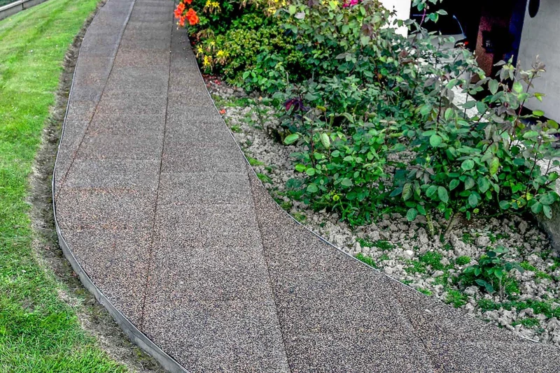 Een gebogen tuinpad van rubbergranulaattegels van WARCO in het kleurendesign Bast en Bark leidt langs het huis door de tuin.