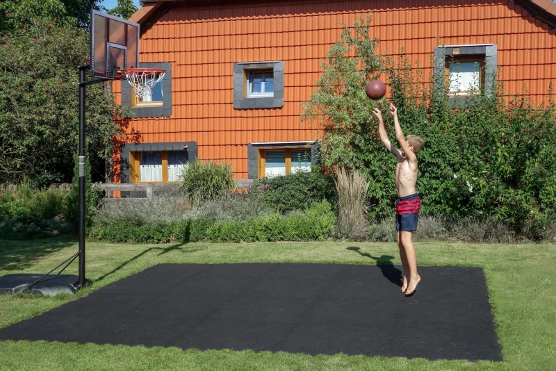 In de tuin bij een vrijstaand huis is een basketbalveld gemaakt van zwarte ballenborden van WARCO. Een jongen gooit de basketbal op de basketbalhoepel, die mobiel is opgesteld.