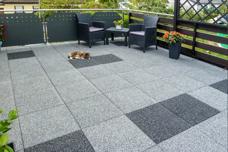 Vierkante terrasplaten in de kleur design Grey en Dark Grey Granite in het formaat 50 × 50 cm zijn gelegd als vloer op een dakterras op een gebouw of garage. In een hoek voor de reling is een zithoek waar een hond ontspannen voor ligt te dutten.
