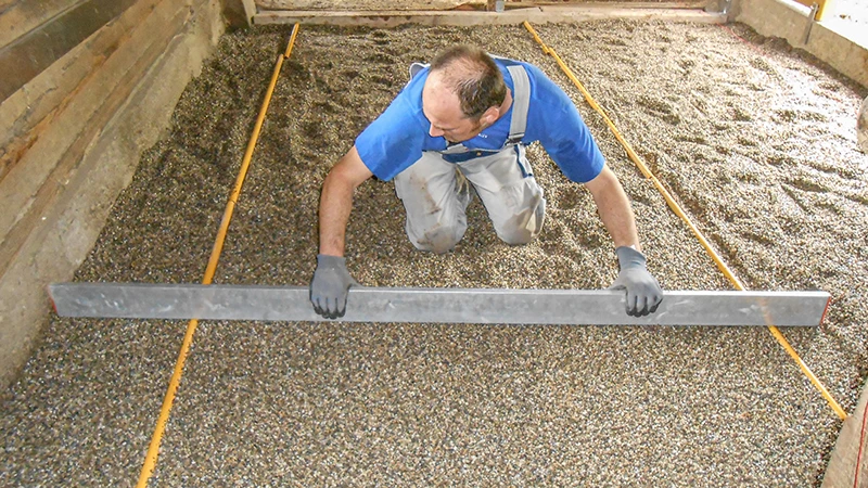Aanbrengen van een nieuwe bodem in een paardenstal: Een man trekt een splitlaag vlak met een rei. De split dient als bedding voor het leggen van een stalvloer van rubbergranulaat. De ruimte wordt aan de zijkanten begrensd door houten of betonnen wanden. Het werk wordt zorgvuldig en nauwkeurig uitgevoerd – typisch voor een correcte opbouw van een paardvriendelijke stalbodem.