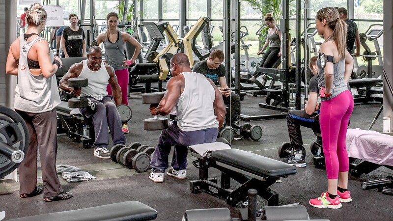 In een LibertyGym fitnessruimte trainen mensen met halters voor een spiegelwand. Verschillende gewichtsstations zijn opgesteld op WARCO fitnessmatten.