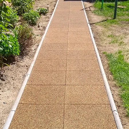 Een recht tuinpad van vierkante rubbertegels met zijafwerking van boordstenen loopt door het perk. De bruine tegels van rubbergranulaat vormen een onderhoudsarme, weerbestendige en antislip ondergrond. Het pad zorgt voor schoon en veilig lopen bij elk weer en scheidt de beplante perken duidelijk van het gazon. Ideaal voor tuinen, erven en toegangen.