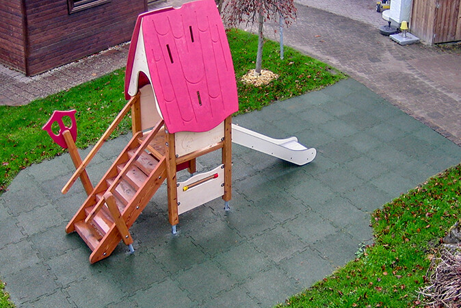 Zicht van bovenaf op een houten speelhuisje met trap en glijbaan. Het puntdak in de stijl van een heksenhuisje is roze. Het speelhuisje staat op schokabsorberende speelvloeren, namelijk op groene valbeschermingsplaten met een puzzelrand van WARCO.