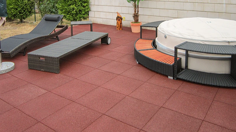 Een ruime buitenruimte met rode WARCO-terrastegels van PU-gebonden rubbergranulaat, die zorgen voor een comfortabele loopervaring en duurzame stevigheid. De plek is ingericht met gezellige tuinmeubels en een whirlpool die naadloos in het geheel past. Het elastische oppervlak dempt voetstappen, is weerbestendig en eenvoudig te onderhouden. Dankzij de slipvaste textuur biedt het een aantrekkelijke oplossing voor stijlvol ontspannen in de buitenlucht. Ideaal voor moderne terrassen die functionaliteit en comfort combineren.