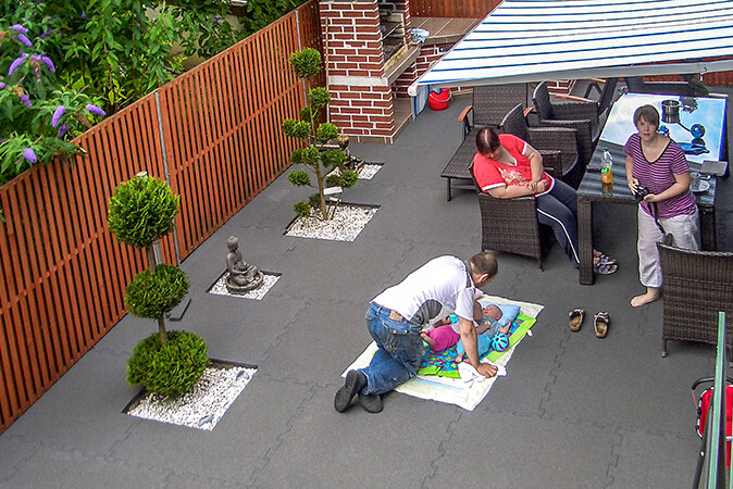 Een gezin geniet van hun vrije tijd op een terras dat is ontworpen als een oase van welzijn met grijze terrasplanken van WARCO. De baby ligt op een kinderdeken op de terrasplanken
