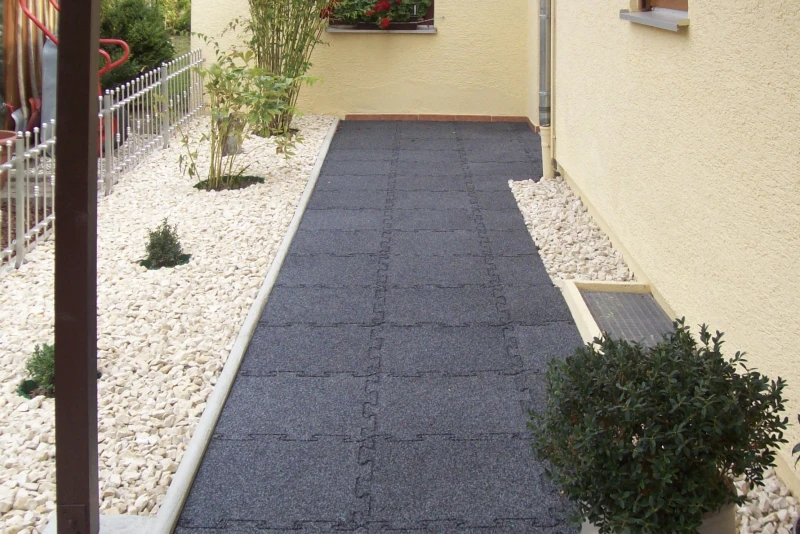 Het voetpad loopt door de tuin aan de zijkant van het huis naar de voordeur. Op de stoep liggen zwarte rubbergranulaatplaten. De tuin is aangelegd als grindtuin, een modieuze en anti-levens slechte gewoonte.